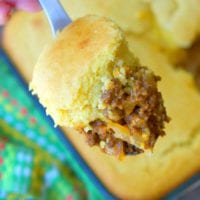 A hand holding a spoonful of cornbread sloppy joe casserole, brimming with a savory ground beef mixture, hovers over a pan of golden cornbread.