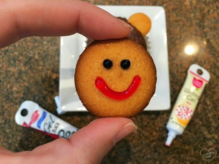 A hand holding a cookie adorned with a red smiley face and black eyes, with icing tubes and another snack in the background, evokes the playful charm of monkey cupcakes at a whimsical bake sale.