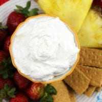 A bowl of creamy pina colada dip sits enticingly surrounded by sliced pineapple, strawberries, and graham crackers on a plate.