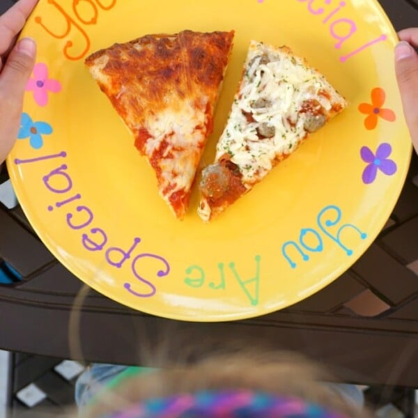 A person proudly holds their DIY "You Are Special" plate in vibrant yellow, showcasing two delicious slices of pizza—one classic cheese and the other loaded with toppings.