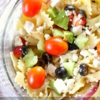 A vibrant Mediterranean pasta salad features a close-up of bowtie pasta adorned with cherry tomatoes, crisp cucumbers, briny olives, feta cheese, and fresh green peppers.