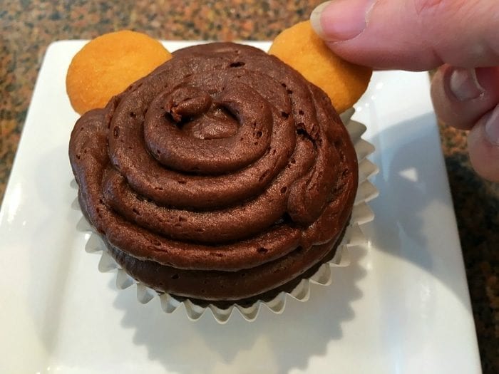 A chocolate cupcake with swirl frosting, crowned by two small vanilla cookies. A hand is playfully placing one cookie, reminiscent of a curious monkey cupcakes design.