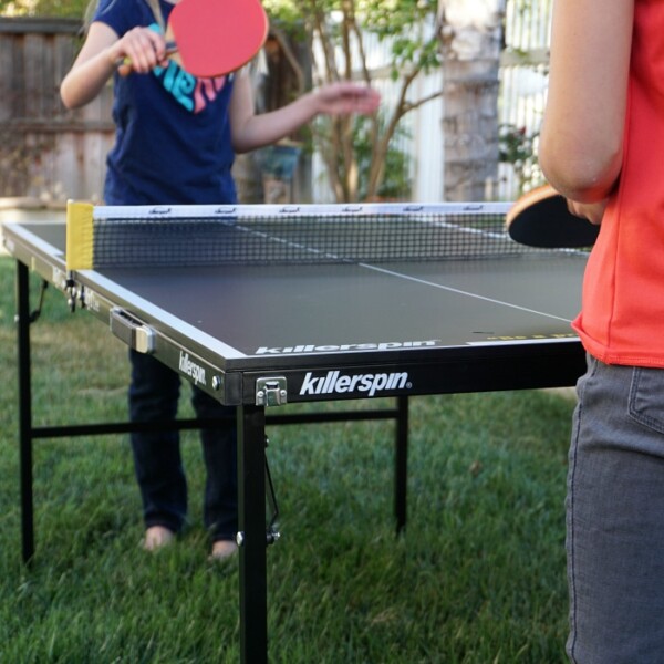 Two people enjoying a lively game of table tennis on a Killerspin mini ping pong table outdoors. The person in the foreground grips a paddle, ready to strike, while their opponent in the background prepares to hit the ball with precision and flair.