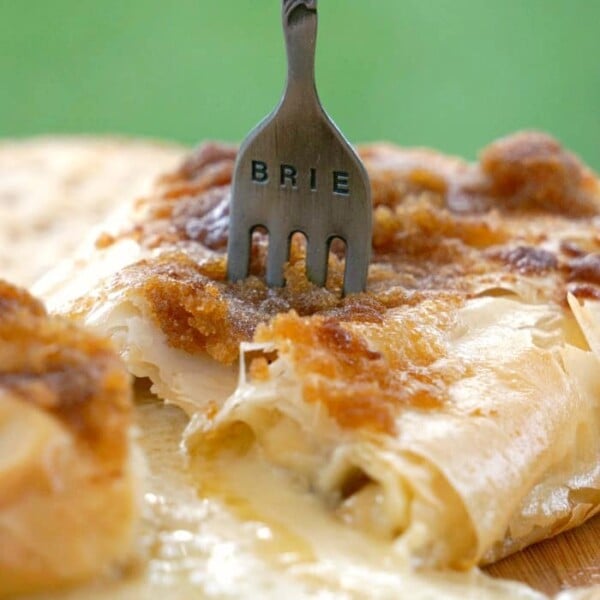 A fork labeled BRIE in melted brie cheese with a flaky phyllo dough crust rests on a wooden board.