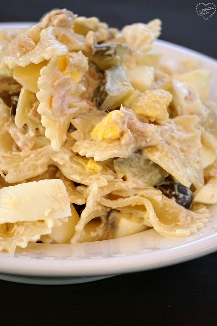 A close-up of a plate containing farfalle pasta mixed with a creamy sauce, reminiscent of a tuna macaroni salad recipe, yellow chunks, and green vegetables. The pasta is arranged in a casual heap on a white dish.