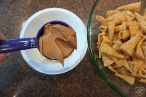 A measuring cup filled with peanut butter is held above a white container, ready to create some unicorn horn muddy buddies. Next to it, a green bowl holds a pile of rolled corn snacks, resting on the brown countertop in perfect harmony.