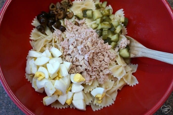 The red bowl holds a vibrant tuna macaroni salad recipe, featuring cooked bowtie pasta, chopped hard-boiled eggs, diced pickles, and sliced olives. A wooden spoon is poised in the bowl, ready for mixing.