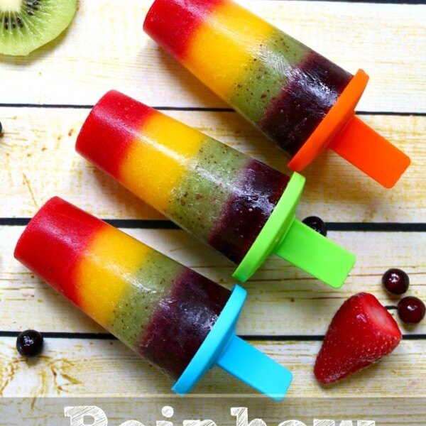 Three vibrant rainbow popsicles rest on a wooden surface, surrounded by an array of fresh fruit slices.