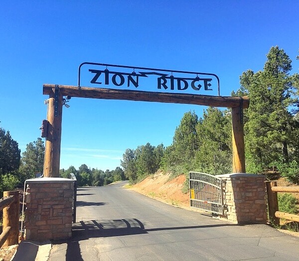 Entrance gate labeled Zion Ridge with a paved road surrounded by trees, leading to the inviting Zion Ponderosa cabins nestled amidst the natural beauty.