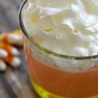 A glass of candy corn parfait, featuring layers of vibrant candy corn pudding topped with a dollop of fluffy whipped cream, sits enticingly. In the backdrop, scattered candy corn pieces add to the festive charm.