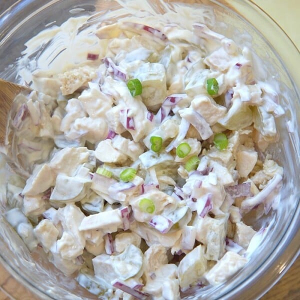 A glass bowl brimming with creamy rotisserie chicken salad, featuring diced onions, peas, and a medley of flavors sits enticingly on a wooden board.