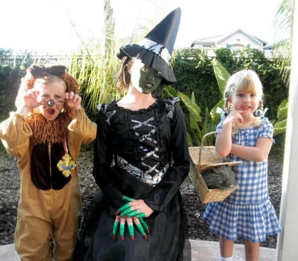 Three children, showcasing a whimsical group Halloween costume idea, are dressed as a lion, a witch, and Dorothy from The Wizard of Oz, standing cheerfully in a garden.