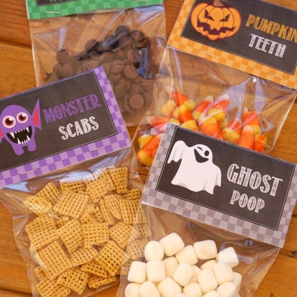 Four labeled Halloween goodie bags of snacks are displayed on a wooden surface. The bags contain: chocolate chips labeled Witches Warts, candy corn labeled Pumpkin Teeth, Chex cereal labeled Monster Scabs, and marshmallows labeled Ghost Poop.