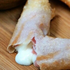 A close-up of a crispy air fryer ham and cheese egg roll on a wooden surface, with melted cheese oozing out from a break in the pastry.