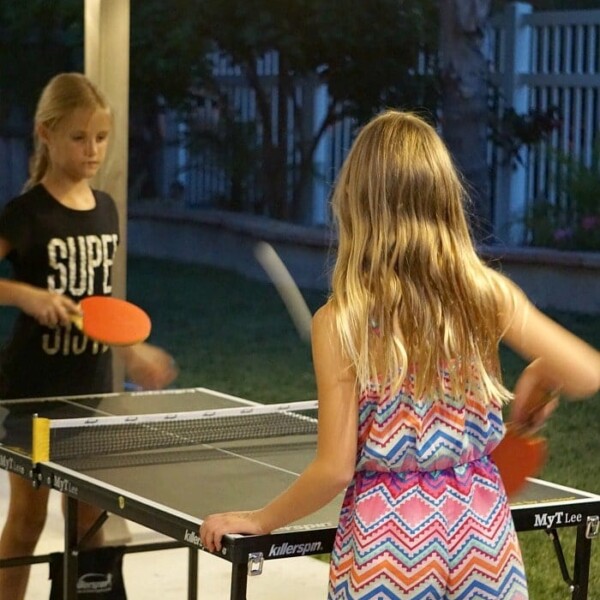 Two girls enjoying a game of ping pong under twinkling string lights in the evening, offering a wonderful way to plug into your kids' world and create lasting memories.