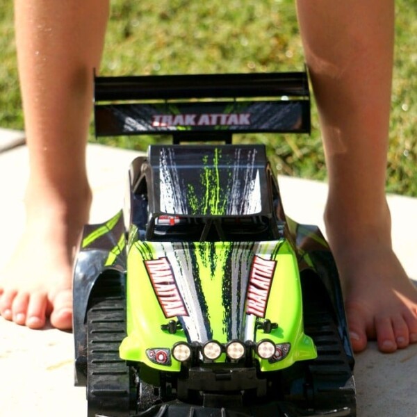 Child's feet beside a green and black RC Trak Attak toy race car on a sunny pavement.