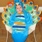 A baby, dressed in a colorful peacock costume with bright feathers, seated indoors, brings joy to family milestone celebrations.