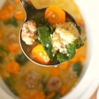 A ladle of easy Italian wedding soup with meatballs, carrots, spinach, and couscous hovers over a bowl.