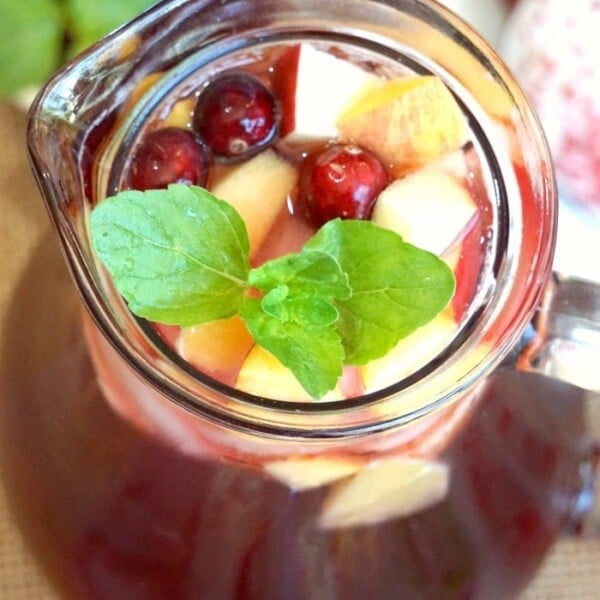 A glass pitcher of sparkling cranberry sangria brimming with apple cubes, fresh cranberries, and a mint garnish.