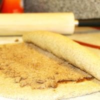 A delicious dough is being skillfully rolled with cinnamon and sugar, hinting at the creation of irresistible gingerbread cinnamon rolls, with a trusty rolling pin in the background.