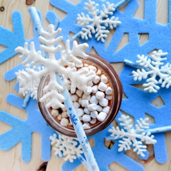 Hot chocolate with marshmallows, surrounded by snowflake decorations, features a blue straw and delightful chocolate snowflake stirrers on a wooden surface.