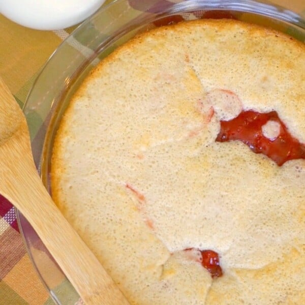 A delectable upside down cherry pie in a glass dish showcases its visible red filling beneath a golden crust. A wooden spatula rests on a plaid cloth, while a glass of milk stands invitingly in the background.