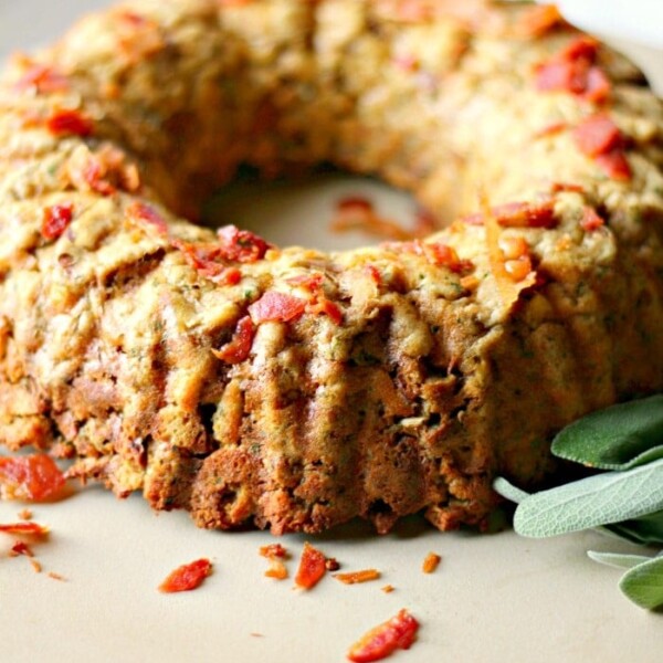 A bundt-shaped meatloaf, crafted in a bundt pan, is adorned with small pieces of bacon and rests on a beige plate. A few green leaves are thoughtfully placed beside it.