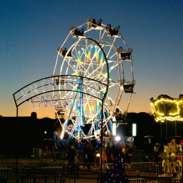 The Ferris wheel, nestled in the grounds of the Fairmont Scottsdale Princess at Christmas, is illuminated at dusk with decorative lights, casting a magical glow as people gather below.