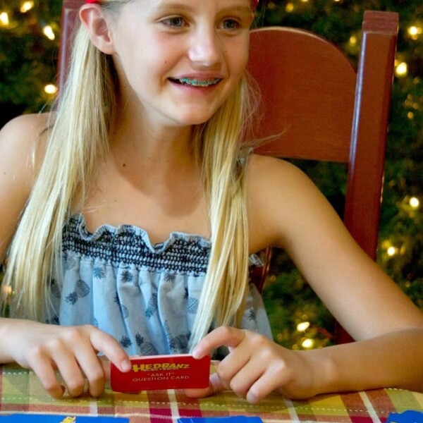 Smiling under holiday lights, a girl enjoys a family game night, playing with the Hedbanz Electronic board game. Her red headband with a card adds to the cheerful atmosphere.