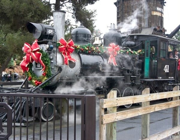 A steam locomotive at Knott's Berry Farm, adorned with holiday wreaths and bows, emits wisps of steam in a festive park setting, capturing the magic of Christmas.