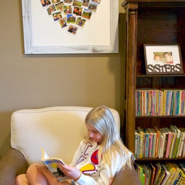 A girl is reading in an armchair beside a bookshelf, with a heart-shaped photo collage and SISTERS picture above, both cherished minted personalized gifts.