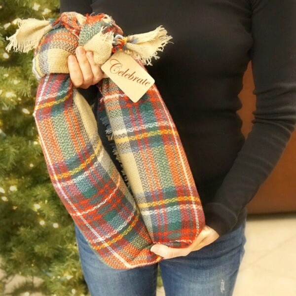 A person in a black shirt and jeans shows how to gift wine bottles in a scarf, holding two bottles wrapped in colorful plaid fabric with frayed edges. The bottles are elegantly tied with a tag reading “Celebrate.” A decorated Christmas tree is partially visible in the background.