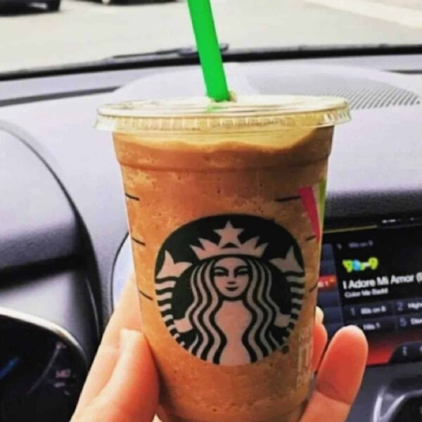 A hand holds a Starbucks Frappuccino with a green straw inside the car, prominently displaying the Starbucks logo. As cars and greenery whizz by through the windshield, one might ponder how to get free Starbucks gift cards to relish such moments more often.