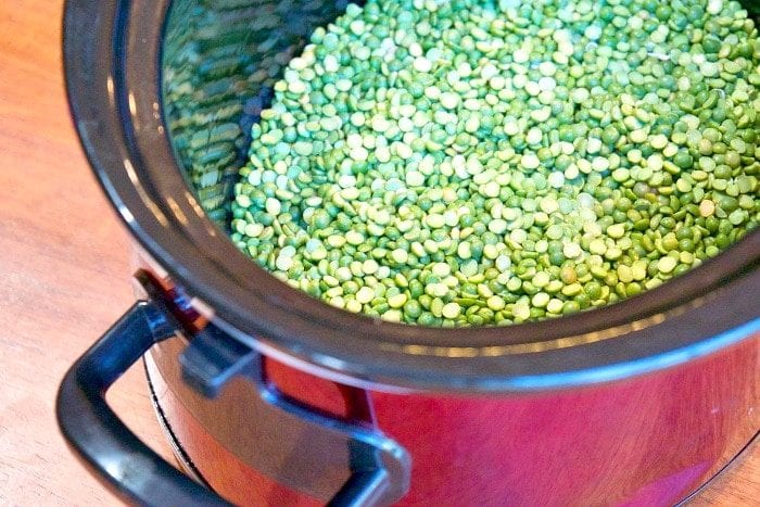 A slow cooker brimming with uncooked green split peas and savory ham rests on a wooden surface, ready to become a hearty split pea soup.
