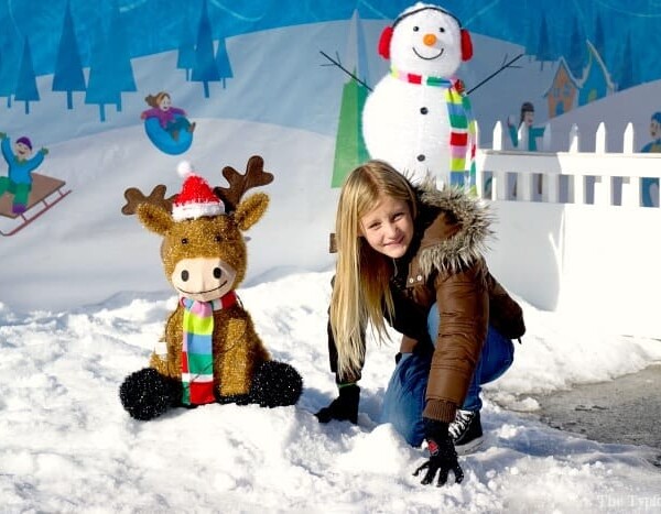 Child in winter coat plays in snow with reindeer toy and snowman backdrop at Winter Wonderfest, bringing the joy of holiday discovery to life.