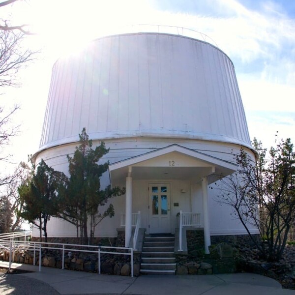 The large white Lowell Observatory building, with its iconic domed roof, is nestled among trees under a sunny sky.