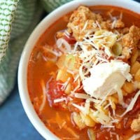 A comforting bowl of instant pot lasagna soup with crumbled meat, diced tomatoes, cheese, and a dollop of cream sits invitingly next to a striped cloth.