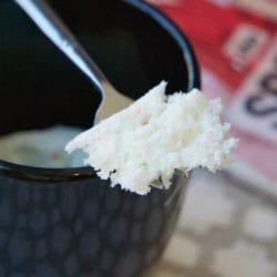 A spoon lifts a textured, angel food cake in a mug over a sleek black cup, with a blurred magazine softly visible in the background.