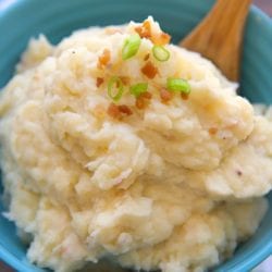 A bowl of pressure cooker mashed potatoes topped with chopped green onions and bacon bits, accompanied by a wooden spoon.