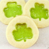 Learn how to make stained glass cookies with three delightful treats featuring green shamrock-shaped translucent centers, elegantly displayed on a light surface.