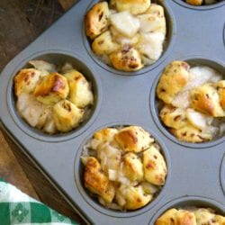 Muffin tin filled with homemade apple pie monkey bread muffins, golden brown and topped with chunks of apple. A green and white checkered cloth is nearby, enhancing the cozy feel.