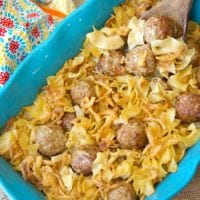 A blue baking dish brimming with pasta and an easy Swedish meatball casserole, topped with a creamy sauce. Baked to perfection, it rests beside a wooden spoon. A colorful patterned cloth adds a touch of warmth in the background.