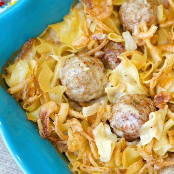 A blue dish holds an easy Swedish meatball casserole, brimming with savory meatballs, egg noodles, crispy fried onions, and a creamy sauce. The ensemble rests on a textured, light-colored surface.