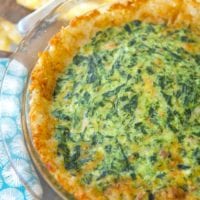 A tater tot spinach quiche sits invitingly in a glass pie dish on a wooden table, surrounded by a cheerful yellow cloth and a vibrant blue napkin.