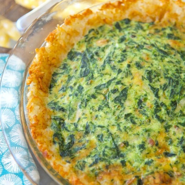 A tater tot spinach quiche sits invitingly in a glass pie dish on a wooden table, surrounded by a cheerful yellow cloth and a vibrant blue napkin.