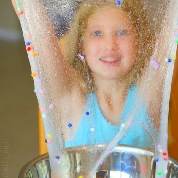 A child in a blue tank top is playing with clear slime containing colorful beads, reminiscent of an easy baking soda slime recipe. The slime is stretched above a stainless steel bowl on the table.