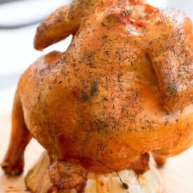 A whole roasted chicken seasoned with herbs sits upright on a stand, reminiscent of a classic beer can chicken cooked on a charcoal grill. Displayed on a wooden cutting board, the bird is golden brown and cooked to perfection, with visible spices adorning its crispy skin.