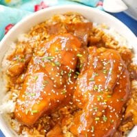 A bowl of rice topped with crockpot teriyaki chicken pieces is garnished with sesame seeds and chopped greens. The dish is served on a blue surface with a floral-patterned cloth nearby.