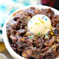 A close-up of a chocolate cake in a white bowl, reminiscent of rich crockpot brownie cookies, topped with a scoop of vanilla ice cream and colorful sprinkles. The background features a blue and white patterned cloth.