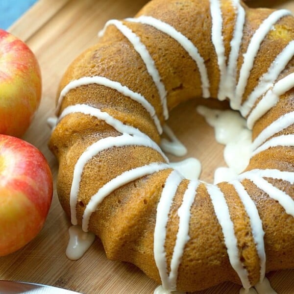 A bundt cake with white icing drizzled on top sits on a wooden cutting board. Made in an Instant Pot, this pumpkin apple creation is accompanied by two apples and a small knife. A colorful cloth with a floral pattern adds charm in the background.